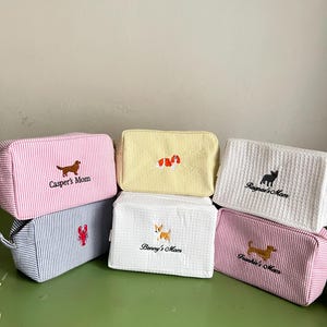 K&ouml;nnte beinhalten: Eine Sammlung von sechs rechteckigen Kosmetiktaschen mit gestickten Hunde-Designs und Namen. Die Taschen weisen verschiedene Farben und Muster auf, darunter rosa und wei&szlig;e Streifen, Gelb, wei&szlig;e Waffel und blau-wei&szlig;e Streifen.
