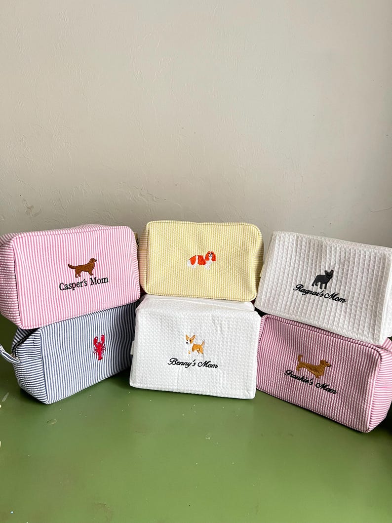 K&ouml;nnte beinhalten: Eine Sammlung von sechs rechteckigen Kosmetiktaschen mit gestickten Hunde-Designs und Namen. Die Taschen weisen verschiedene Farben und Muster auf, darunter rosa und wei&szlig;e Streifen, Gelb, wei&szlig;e Waffel und blau-wei&szlig;e Streifen.