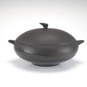 May include: A matte black lidded serving bowl with a smooth, rounded silhouette. The bowl features two small, curved side handles and a matching domed lid with a central finial handle. The design is uniform in colour and has a clean, curved shape.