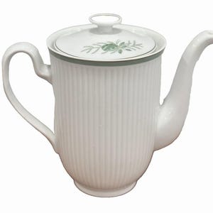 Puede incluir: Tetera de cerámica blanca con tapa. La tetera tiene un diseño acanalado y un asa curva. La tapa presenta un estampado floral verde y un borde verde. El pico es curvo.