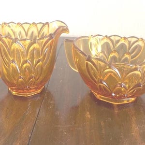 May include: Amber-colored glass creamer and sugar bowl set. The pieces feature a floral design and are set on a wooden surface. The creamer has a handle and spout, while the sugar bowl has two handles.