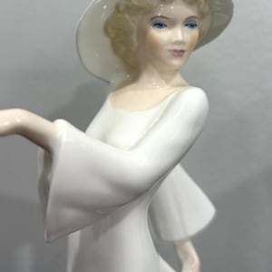 May include: A porcelain figurine of a woman in a white dress and hat. The figure has blonde hair, blue eyes, and is posed with one arm extended. The dress has flowing sleeves and a long skirt. The figurine is a decorative item.