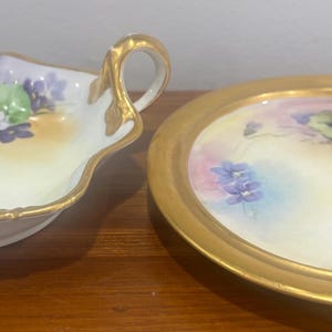 Puede incluir: Dos fuentes antiguas de porcelana. Una es una fuente con asa y borde festoneado con un diseño floral en morado, verde y amarillo, y ribete dorado. La otra es una fuente redonda con borde dorado y un diseño floral similar.