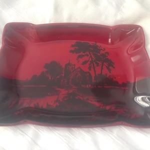 May include: A rectangular, red glass ashtray with a black landscape scene. The scene depicts a church, trees, and a body of water. The ashtray has rounded corners and a slightly raised edge.