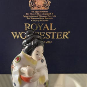 Puede incluir: Una figura de porcelana Royal Worcester de una persona con el cabello negro recogido en un moño, vistiendo una túnica blanca decorada con patrones circulares coloridos. La figura está frente a una caja azul oscuro con el texto Royal Worcester.