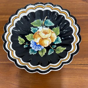 May include: A round ceramic bowl with a scalloped edge, featuring a glossy black interior decorated with a central hand-painted floral motif in yellow, blue, and green. The rim is accented with concentric bands of white, orange, and black.