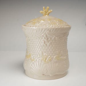 May include: A cream-colored ceramic canister with a lid featuring a textured surface. The body displays embossed patterns of seashells, coral, and wave motifs. The lid is topped with a small, star-shaped finial with a glossy, pearlescent finish.