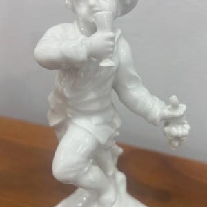 Może przedstawiać: Biała porcelanowa figurka młodego człowieka trzymającego kielich i kiść winogron. Postać ma na sobie kapelusz i koszulę z długim rękawem. Figurka jest zamontowana na kwadratowej podstawie.