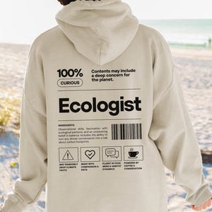 Può includere: Una felpa con cappuccio beige chiaro con la parola "Ecologist" stampata a grandi lettere nere sul retro. La felpa con cappuccio presenta anche il testo "100% Curious" e un elenco di "ingredienti" con icone. Lo sfondo è una scena di spiaggia.