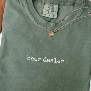 Peut inclure: Un t-shirt à manches courtes vert mousse avec une encolure ronde et le texte "beer dealer" brodé en blanc sur la poitrine. L'étiquette intérieure indique "Comfort Colors" et une taille small. Un collier doré fin repose sur le t-shirt.