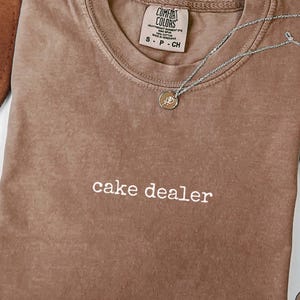 以下が含まれることがあります： 胸元に白い糸で「cake dealer」と刺繍された茶色の半袖Tシャツ。Tシャツの内側の襟元にはComfort Colorsのブランドラベルが付いており、繊細なシルバーカラーのチェーンネックレスが添えられています。