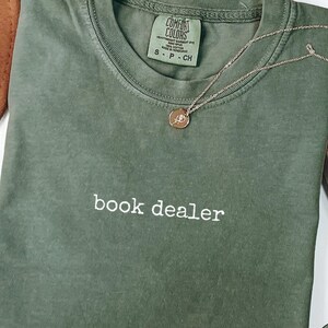 May include: A moss green short-sleeved t-shirt with the text "book dealer" embroidered in white thread on the chest. The shirt features a crew neckline and a Comfort Colors brand label on the inside collar, accompanied by a delicate gold necklace.