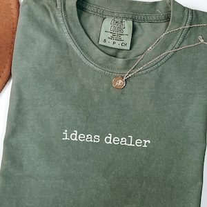以下が含まれることがあります： モスグリーンの半袖Tシャツで、クルーネック仕様。胸元には白い糸で「ideas dealer」の文字が刺繍されています。首の内側にはComfort Colorsのブランドラベルがあり、繊細なゴールドトーンのチェーンネックレスが添えられています。