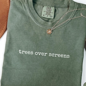Può includere: Una t-shirt a maniche corte color verde muschio con scollo rotondo e la scritta bianca "trees over screens" stampata sul petto. La maglietta presenta un'etichetta del marchio Comfort Colors sul collo interno, che indica la taglia S, in cotone.