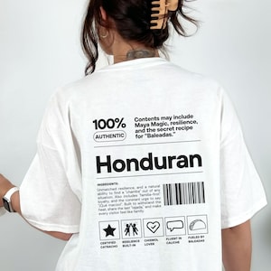 May include: White t-shirt with the word "Honduran" in bold black letters. The shirt includes text about ingredients and a list of icons. The shirt also has the text "100% Authentic" and "Contents may include Maya Magic, resilience, and the secret recipe for Baleadas."
