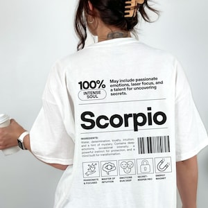 Pode incluir: Camiseta branca com a palavra "Scorpio" em letras pretas grandes. A camiseta apresenta texto sobre os tra&ccedil;os de Escorpi&atilde;o, incluindo "Intense Soul" e "Passionate & Focused". A camiseta tamb&eacute;m tem um design de c&oacute;digo de barras.
