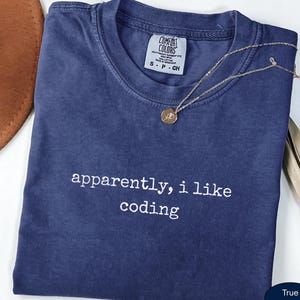 May include: A navy blue short-sleeved t-shirt with a crew neckline and the white embroidered text "apparently, i like coding" centered on the chest. The shirt features a Comfort Colors brand label at the inner neck and a thin gold-toned necklace.