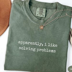 Op de afbeelding: Een mosgroen t-shirt met ronde hals en de witte tekst "apparently, i like solving problems" op de voorzijde. Het shirt is voorzien van een Comfort Colors merklabel bij de binnenhals en wordt gecombineerd met een fijne goudkleurige ketting.