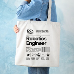 Op de afbeelding: Een witte canvas draagtas met de woorden "Robotics Engineer" in vette zwarte letters. De tas heeft extra tekst, waaronder "100% Analytical Thinking" en een ingrediëntenlijst. De tas wordt door een persoon vastgehouden.