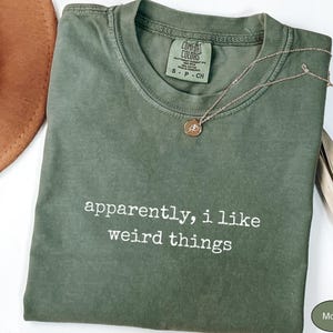 May include: A moss green crew neck t-shirt with the white text "apparently, i like weird things" printed on the front. The shirt features a Comfort Colors brand label at the inner neckline and is paired with a delicate gold-toned chain necklace.