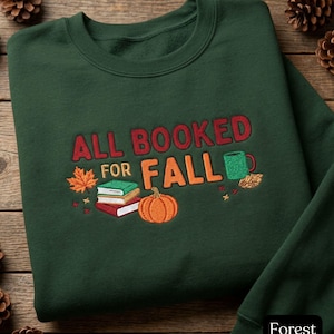 Puede incluir: Una sudadera de cuello redondo en color verde bosque con un bordado que dice ALL BOOKED FOR FALL. El diseño incluye una pila de tres libros, una calabaza naranja, una taza verde y una hoja de arce naranja con pequeñas estrellas decorativas.