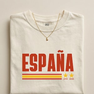 May include: An ivory crew neck t-shirt featuring the red text ESPAÑA, a horizontal red and yellow striped graphic, and two yellow stars with the years 2010 and 2026 printed below them. A thin gold-toned chain necklace rests on the neckline.