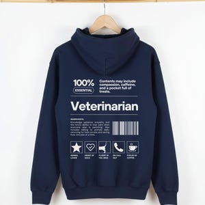 May include: Navy blue hoodie with the word "Veterinarian" in white block letters. The back of the hoodie has text that reads "100% Essential" and lists ingredients like compassion, caffeine, and treats. Icons of a star, heart, dog, phone, and coffee cup are also present.