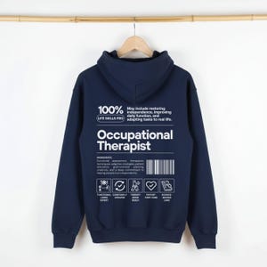 May include: A navy blue hooded sweatshirt featuring white text and graphics on the back. The design includes the words Occupational Therapist, a barcode, and icons labeled Functional Living Expert, Constantly Updated, Therapy Mode Ready, Patient First Care, and Science-Backed Care.