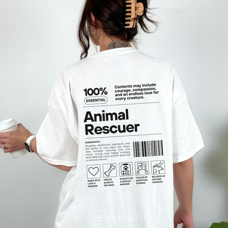 Op de afbeelding: Wit T-shirt met de tekst "Animal Rescuer" in zwart. Het shirt bevat een lijst met ingredi&euml;nten en pictogrammen met betrekking tot dierenredding. De tekst "100% Essential" staat ook op het shirt.