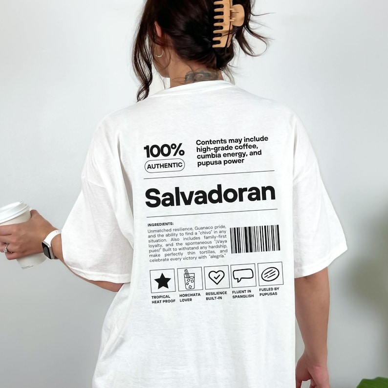 Pu&ograve; includere: T-shirt bianca con la scritta "Salvadoran" a grandi lettere nere. La maglietta presenta il testo "100% Autentico" e "Il contenuto pu&ograve; includere caff&egrave; di alta qualit&agrave;, energia cumbia e pupusa power". Ulteriori testi e icone sono stampati in basso.