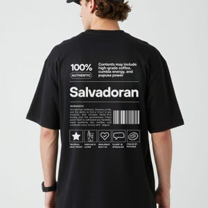 Pu&ograve; includere: T-shirt nero con testo e grafica bianchi. La maglietta presenta la parola "Salvadoran" e il testo "100% Authentic". Il testo aggiuntivo include ingredienti e icone. &Egrave; un capo casual.