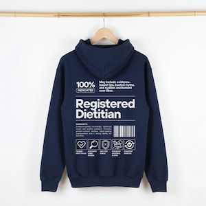 Pu&ograve; includere: Felpa con cappuccio blu navy con la scritta "Registered Dietitian" in bianco. Il retro della felpa presenta una grafica con le parole "100% Dedicated" e un elenco di ingredienti relativi alla nutrizione. La felpa &egrave; appesa a una gruccia di legno.