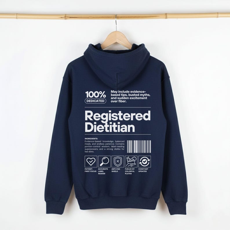 Pu&ograve; includere: Felpa con cappuccio blu navy con la scritta "Registered Dietitian" in bianco. Il retro della felpa presenta una grafica con le parole "100% Dedicated" e un elenco di ingredienti relativi alla nutrizione. La felpa &egrave; appesa a una gruccia di legno.