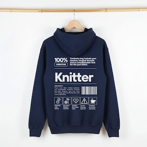 Op de afbeelding: Een marineblauwe hoodie met het woord "Knitter" in grote witte letters. De achterkant van de hoodie heeft tekst over breien, waaronder "100% Creative" en een ingredi&euml;ntenlijst. De hoodie hangt aan een houten hanger.