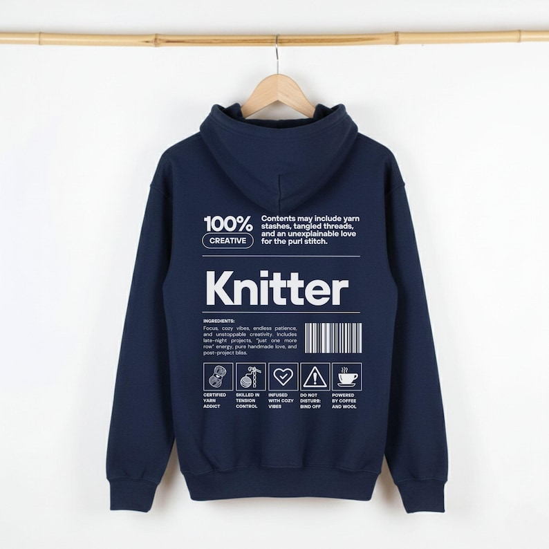 Op de afbeelding: Een marineblauwe hoodie met het woord "Knitter" in grote witte letters. De achterkant van de hoodie heeft tekst over breien, waaronder "100% Creative" en een ingredi&euml;ntenlijst. De hoodie hangt aan een houten hanger.