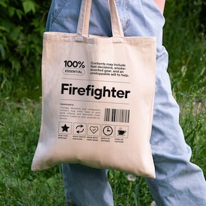 Könnte beinhalten: Beige Canvas-Tragetasche mit dem Wort "Firefighter" in schwarzer Schrift. Die Tasche enthält Text und Symbole. Sie wird von einer Person in blauen Jeans vor einem grünen Hintergrund getragen.
