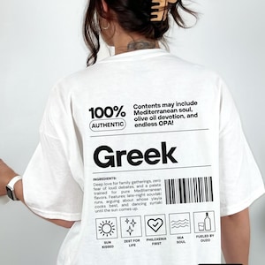 Può includere: T-shirt bianco con una grande grafica nera che recita "Greek". Il design include testo sull'anima mediterranea, l'olio d'oliva e OPA! Il testo aggiuntivo elenca ingredienti e caratteristiche, insieme a icone.