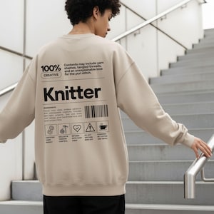Op de afbeelding: Beige sweatshirt met het woord "Knitter" in zwarte letters. De achterkant van de sweatshirt heeft een afbeelding met de tekst "100% Creative" en "Contents may include yarn stashes, tangled threads, and an unexplainable love for the purl stitch."