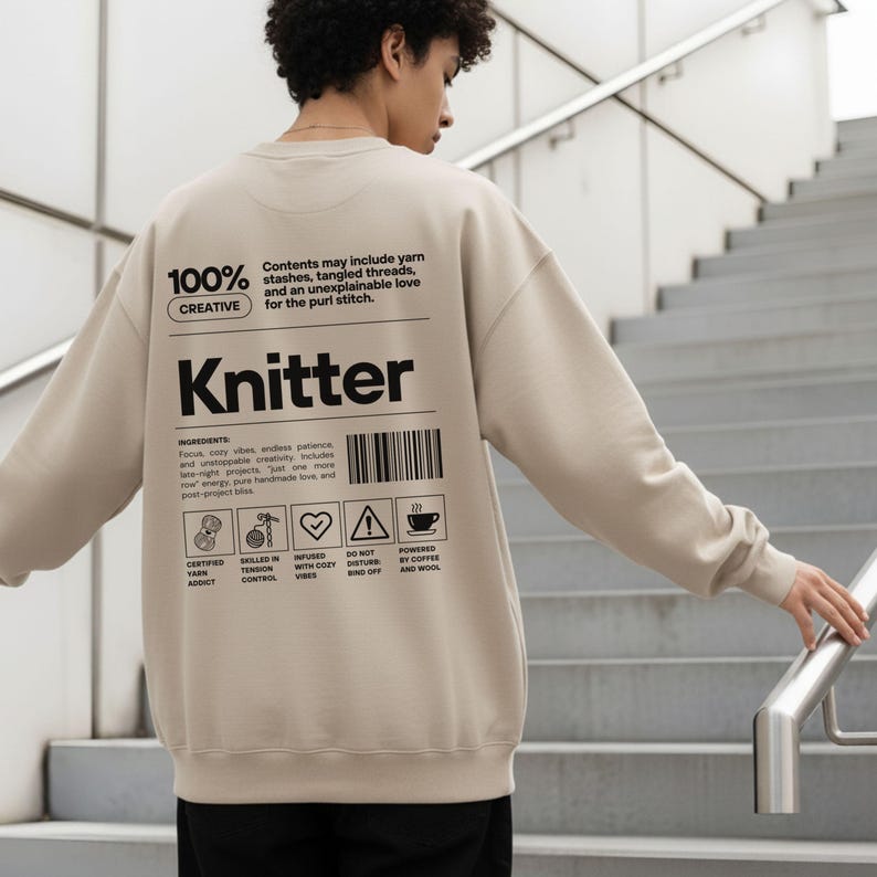 Op de afbeelding: Beige sweatshirt met het woord "Knitter" in zwarte letters. De achterkant van de sweatshirt heeft een afbeelding met de tekst "100% Creative" en "Contents may include yarn stashes, tangled threads, and an unexplainable love for the purl stitch."