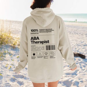 K&ouml;nnte beinhalten: Ein beigefarbener Kapuzenpullover mit dem Text "ABA Therapist" in Schwarz. Auf der R&uuml;ckseite des Kapuzenpullovers befindet sich eine Grafik mit dem Text "100% Supportive" und anderen Texten und Symbolen. Der Kapuzenpullover ist ein l&auml;ssiges Kleidungsst&uuml;ck.