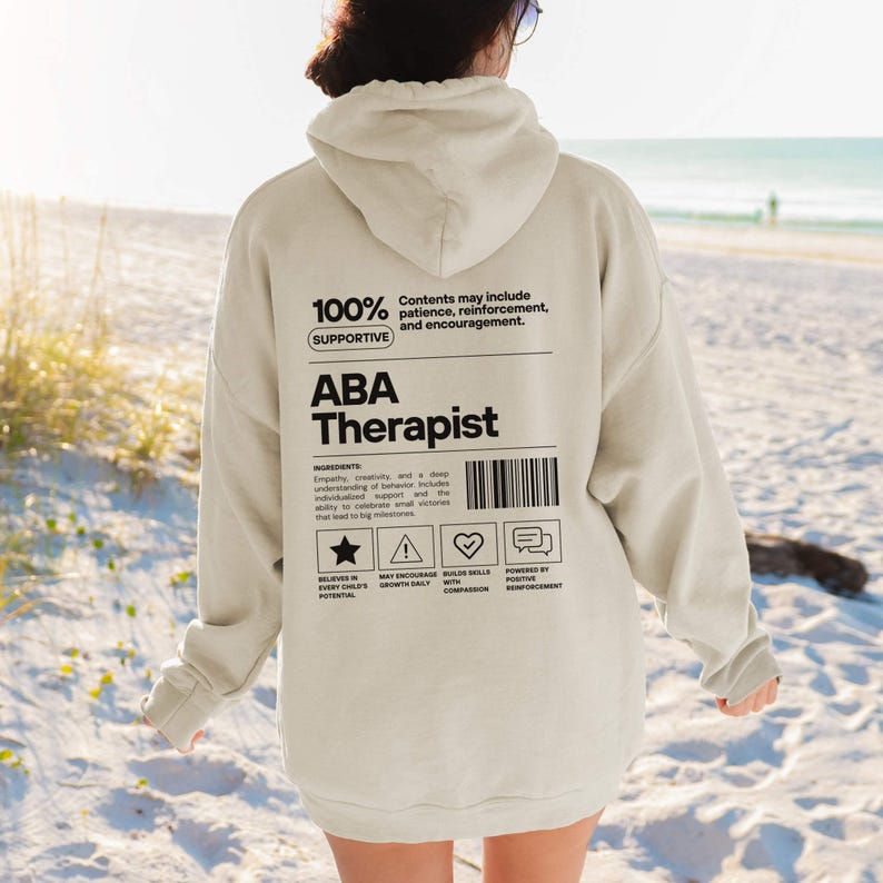 K&ouml;nnte beinhalten: Ein beigefarbener Kapuzenpullover mit dem Text "ABA Therapist" in Schwarz. Auf der R&uuml;ckseite des Kapuzenpullovers befindet sich eine Grafik mit dem Text "100% Supportive" und anderen Texten und Symbolen. Der Kapuzenpullover ist ein l&auml;ssiges Kleidungsst&uuml;ck.