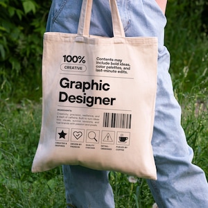 Puede incluir: Bolso tote beige con el texto "Graphic Designer" en letras negras. La bolsa incluye las palabras "100% CREATIVE" y una lista de ingredientes. Tiene un código de barras e iconos de inspiración, pasión, calidad, detalle y café.