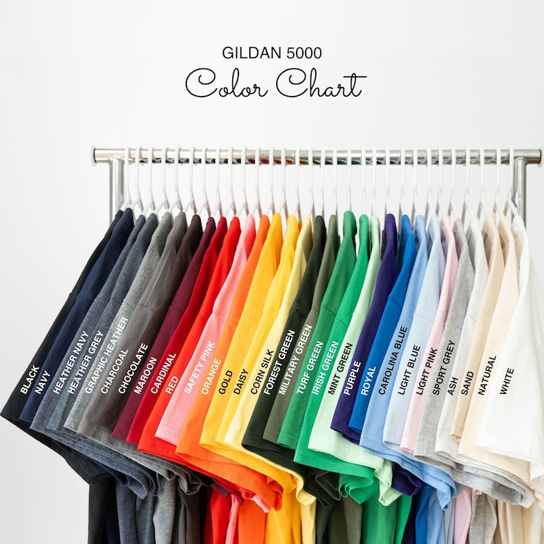 Pode incluir: Uma tabela de cores para camisetas Gildan 5000, mostrando uma variedade de cores, incluindo preto, azul marinho, cinza mesclado, vermelho, laranja, verde, azul, rosa e branco. A tabela &eacute; exibida em um cabideiro de metal com cabides brancos.