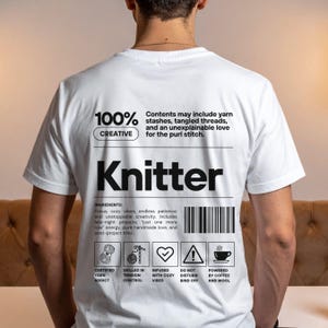 Op de afbeelding: Wit T-shirt met het woord "Knitter" in grote zwarte letters. Het shirt heeft de tekst "100% Creatief" en "Inhoud kan garenvoorraden, verwarde draden en een onverklaarbare liefde voor de averechte steek bevatten."