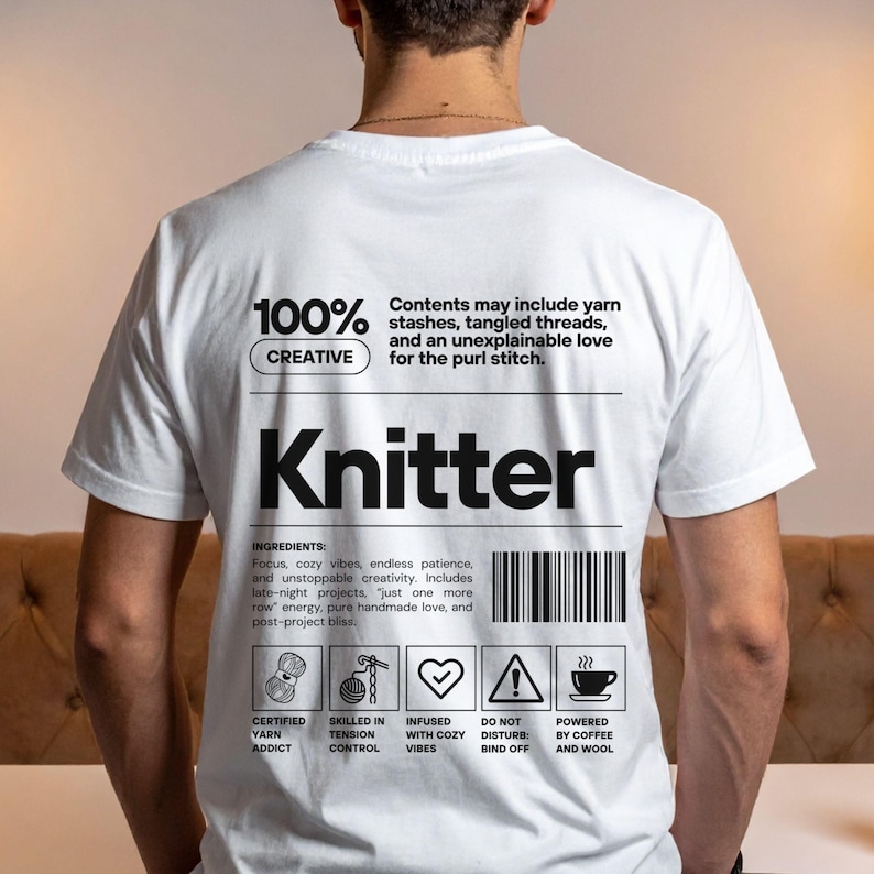 Op de afbeelding: Wit T-shirt met het woord "Knitter" in grote zwarte letters. Het shirt heeft de tekst "100% Creatief" en "Inhoud kan garenvoorraden, verwarde draden en een onverklaarbare liefde voor de averechte steek bevatten."