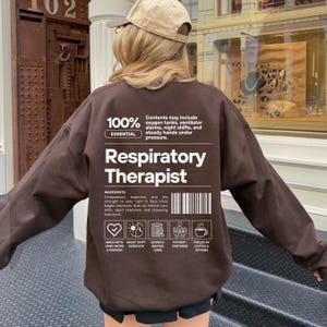 Może przedstawiać: Brązowa bluza z napisem "Respiratory Therapist" w kolorze białym. Bluza zawiera r&oacute;wnież dodatkowy tekst i ikony, w tym "100% Essential" i "Made with Hard Work & Passion."