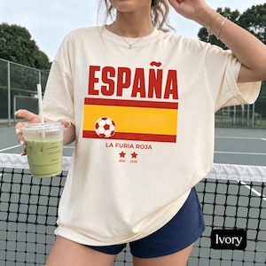 Può includere: Una t-shirt a maniche corte color avorio con la scritta rossa ESPAÑA sopra una grafica a strisce orizzontali rossa e gialla. Un pallone da calcio è centrato sulla striscia. Sotto, il testo LA FURIA ROJA appare con due stelle rosse 2010 e 2026.