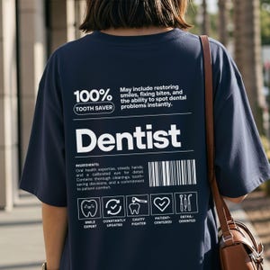 Puede incluir: Camiseta azul marino con la palabra "Dentist" en letras blancas grandes. La camiseta presenta el texto "100% Tooth Saver" y texto adicional que describe los ingredientes. También hay iconos de dientes y herramientas dentales.