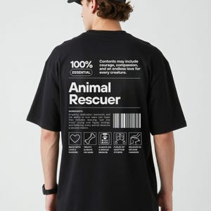 Op de afbeelding: Zwart t-shirt met de witte tekst "Animal Rescuer". Het ontwerp bevat "100% Essential" en een ingredi&euml;ntenlijst. De achterkant van het shirt heeft iconen en tekst over dierenredding.