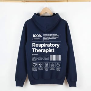 May include: Navy blue hoodie with white text reading "Respiratory Therapist". The back of the hoodie features a list of ingredients and icons. The hoodie is suspended from a wooden hanger.